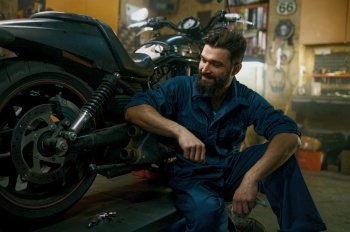 Satisfied smiling professional mechanic feeling glad looking at repaired motorcycle after good work in repair workshop. Satisfied smiling mechanic feeling glad looking at repaired motorcycle