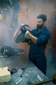 Serious repairman inspecting welded gas tank for motorcycle. Serviceman working at smoky workshop in evening. Serious concentrated repairman inspecting new welded part for motorcycle