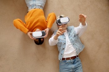 Overhead view on young attractive lesbian woman couple using vr goggles headset lying on floor at home living room. Happy girlfriends watching movie, playing games in virtual reality. Attractive lesbian woman couple using vr goggles headset lying on floor