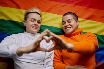 Selective focus on couple with heart sign hands gesture. Young lesbian woman couple lying over rainbow colored flag. Selective focus on couple with heart sign hands gesture