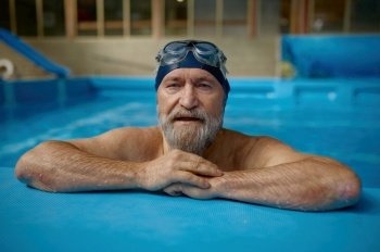 Portrait of mature senior man wearing swimming hat and goggles at pool. Grey-haired bearded male swimmer looking at camera. Concept of sport, active lifestyle and retirement. Portrait of mature senior man wearing swimming hat and goggles at pool