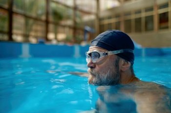 Elderly man swimming in indoor pool enjoying retirement holiday with relaxation. Side view shot of senior male swimmer head in cap and glasses. Elderly man swimming in indoor pool enjoying retirement holiday with relaxation