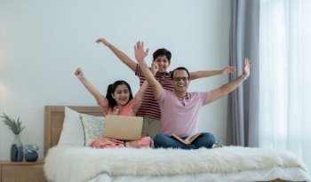 Cheerful indian family teenage boy son and girl with dad have fun and raising hands up together while relaxing and using laptop at home, single father with children sitting on bed at bedroom at home