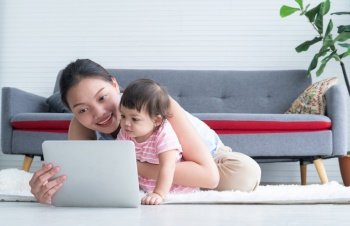 Happy young Asian beautiful mother sitting on floor, working with tablet while holding her cute little Caucasian 7 months newborn baby, taking care infant at home. Child care concept