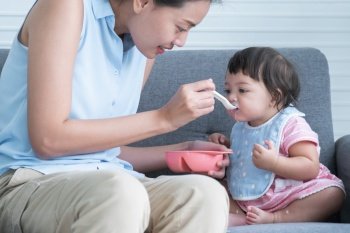 Caucasian Cute 7 month newborn baby girl eating blend mashed food, sitting on sofa with apron at home, young Asian mother holding spoon feeding child, baby mouth mess up with food. Healhy food for kid