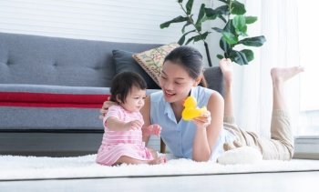 Happy multiracial family, young Asian mother lying on floor, playing with Caucasian newborn 7 months old baby girl at home with yellow duck toy and doll. Education and development children concept