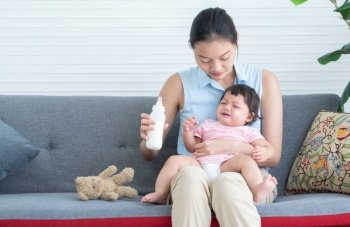 Young beautiful mother holding and feeding milk to cute little Caucasian 7 months newborn baby,  sitting together on sofa at home.Crying infant with unhappy face and deny drinking milk from bottle