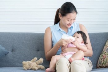 Young Asian beautiful mother smiling, holding and feeding milk from bottle to cute little Caucasian 7 months newborn baby,  sitting together on sofa at home. Child care concept