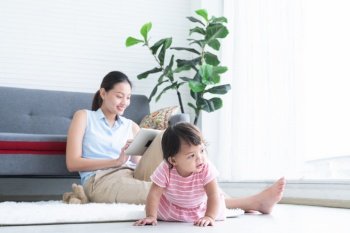 Young Asian beautiful mother sitting on floor, working with tablet while her cute little Caucasian 7 months newborn baby crawling on floor playing near mom at home. Child care concept