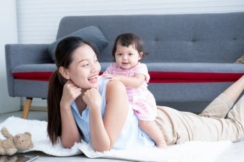 Young Asian mother lying on floor, giving cute 7 month newborn baby girl piggyback ride, enjoying active leisure time. Smiling little infant playing sitting on mom back, having fun together