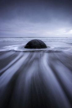 Dramatic streaks of water lead into and splash around this ancient boulder on Moeraki Beach in New Zealand during a blue morning.