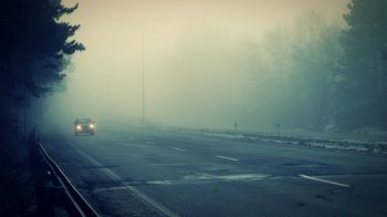 Car traveling on the foggy road with headlights or headlamps on. Low visibility - Dangerous driving of cars in winter in bad weather.