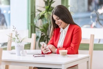 Young attractive smiling businesswoman in red jacket and long hair looking on smart phone while sitting at table in cafe. Employee woman looking at mobile phone in cafe