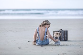 Back view of girl in denim overall shorts petting small Yorkshire Terrier dog while sitting on sand near suitcase on Famara Beach in Lanzarote, Spain. Girl caressing dog on beach