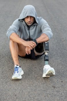 Unhappy middle aged sportsman in gray hoodie with prosthetic leg sitting on asphalt road during fitness workout. Handicapped male athlete sitting on asphalt road