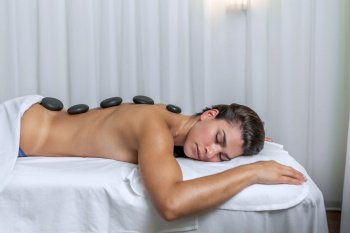 Side view of a young latina woman lying face down in a massage table with spa stones on her back. Concept of spa.. Side view of a young woman lying face down in a massage table with spa stones on her back