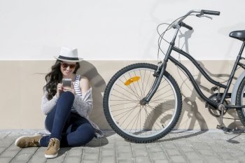 Young cheerful woman in sunglasses and hat sitting nearby bike and using device. Lanzarote, Gran Canaria, Spain. . Young woman with device sitting on ground