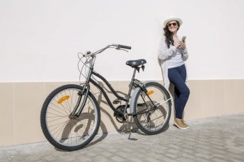 Young smiley woman in hat and sunglasses with bicycle and gadget standing against wall, Gran Canaria, Spain. . Smiley female with device and bicycle