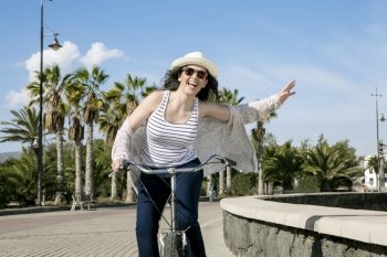 Young cheerful woman in hat and sunglasses bicycling on quay agaist palms of Gran Canaria in Spain. . Young happy woman cycling in Spain