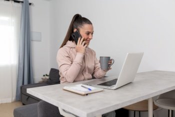 Smiling young female freelancer in casual clothes having phone conversation and drinking cup of hot coffee during break from remote work on laptop at home. Happy woman talking on smartphone during break