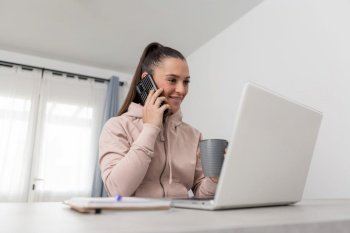 Cheerful young female freelancer in hoodie looking at laptop screen and smiling while talking on smartphone during coffee break from remote work at home. Smiling woman speaking on mobile phone with coffee cup