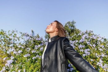 pretty young woman with blonde hair and dressed in a leather jacket, with her eyes closed breathing fresh air in the forest on a sunny day