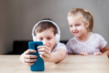 two children playing on the smart phone in headphones. people, children and technology concept - kids with mobile taking selfie or having video call at home.. two happy children playing on the smartphone in headphones. people, children and technology concept