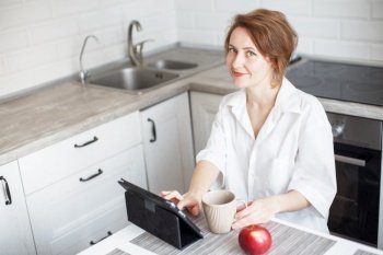 Happy beautiful woman with cup of coffee or tea using laptop in quarantine lockdown in the kitchen at home in the white shirt. Social distancing Self Isolation. Happy beautiful woman with cup of coffee or tea using laptop in quarantine lockdown in the kitchen in the white shirt.