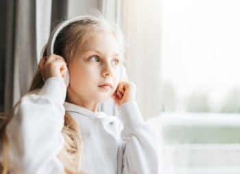 Little girl with headphones indoors at home, sitting on window sill and listening music