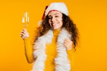 Young cute girl smiling and holding glass of champagne or wine on yellow studio background. Woman in Santa hat. New year mood. High quality photo. Young cute girl smiling and holding glass of champagne or wine on yellow studio background. Woman in Santa hat. New year mood.