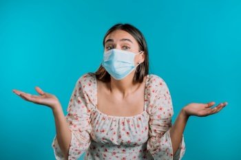 Unsure woman in medical mask shrugs her arms, makes gesture of I don’t know, care, can’t help anything . Girl on blue background.. Unsure woman in medical mask shrugs her arms, makes gesture of I don’t know, care, can’t help anything . Girl on blue background