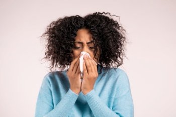 African woman sneezes into tissue. Isolated girl on beige studio background. Lady is sick, has a cold or allergic reaction. Coronavirus, epidemic 2021, illness concept.. African woman sneezes into tissue. Isolated girl on beige studio background. Lady is sick, has a cold or allergic reaction. Coronavirus, epidemic 2021, illness concept