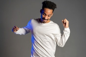 Young very active and energetic african american man in white wear smiling and dancing in good mood on grey background. . Young very active and energetic african american man in white wear smiling and dancing in good mood on grey background