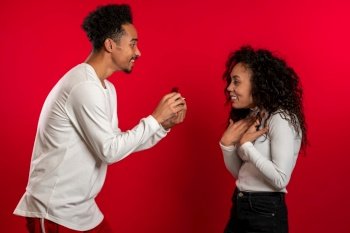 Young couple. African man makes marriage proposal to his lover woman with ring on red studio background. Love, holidays, happiness concept. Young couple. African man makes marriage proposal to his lover woman with ring on red studio background. Love, holidays, happiness concept.