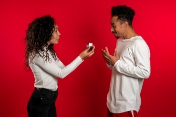 Young couple. African woman makes marriage proposal to her lover man with ring on red studio background. Feminism, equality rights concept.. Young couple. African woman makes marriage proposal to her lover man with ring on red studio background. Feminism, equality rights concept