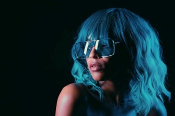 Portrait of alluring blue haired mixed race girl in neon light. Fashion, glamour, model concept. Seductive woman with make-up and transparent glasses posing in dark room at night.. Portrait of alluring blue haired mixed race girl in neon light. Fashion, glamour, model concept. Seductive woman with make-up and transparent glasses posing in dark room at night
