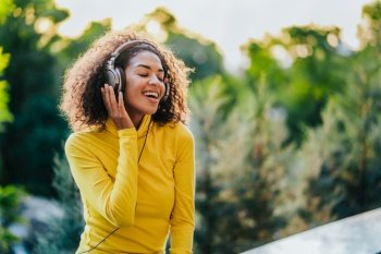 African teenager girl listens to music with headphones in park. Cute woman in yellow, dancing with head, curly hair. Student, freedom, music concept. African teenager girl listens to music with headphones in park. Cute woman in yellow, dancing with head, curly hair. Student, freedom, music concept.