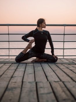 Young brunette woman doing yoga exercises on wooden sea embankment in the morning. Girl in black sports costume. Stretching, practice, healthy lifestyle concept.. Young brunette woman doing yoga exercises on wooden sea embankment in the morning. Girl in black sports costume. Stretching, practice, healthy lifestyle concept