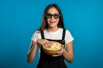 Young asian girl in 3d glasses watching fascinating comedy movie, laughing and eating popcorn on blue studio background. Young asian girl in 3d glasses watching fascinating comedy movie, laughing and eating popcorn on blue studio background.