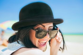 Close up portrait of a woman in blue shirt , sunglasses and summer black bowler trendy hat. Beauty cute girl on a tropical beach sea ocean shore with large stones. Outdoor summer lifestyle.. Portrait of smiling beautiful young woman in black hat and retro sunglasses on the beach