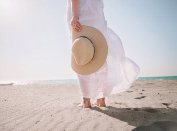 Beautiful caucasian female girl holding hat on the sea beach. Woman wearing in long white dress, standing on sand. Windy weather. Travel concept.. Beautiful caucasian girl holding hat on the sea beach. Woman wearing in long white dress, standing on sand. Windy weather. Travel concept.