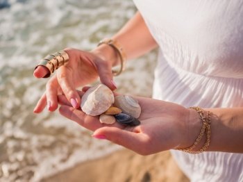 Hand holding sea shells and pumice stones found washed on rocky beach.. Hand holding sea shells and pumice stones found washed on rocky beach. close up hands with boho gypsy accessories. Woman standing on the beach at summer near sea.