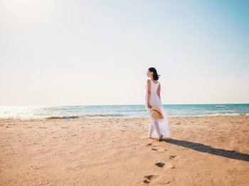 Woman wearing white maxi long dress walking barefoot on the sea shore with hat in hands. Bohemian clothing style. High quality photo. Woman wearing white maxi long dress walking barefoot on the sea shore with hat in hands. Bohemian clothing style.