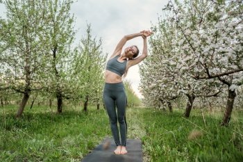 Young woman doing yoga exercise in apple blossoming garden, natural background. Concentrated girl training at summer outdoors. Pastel colors, unity with nature, balance, lifestyle concept. High photo. Young woman doing yoga exercise in apple blossoming garden, natural background. Concentrated girl training at summer outdoors. Pastel colors, unity with nature, balance, lifestyle concept.
