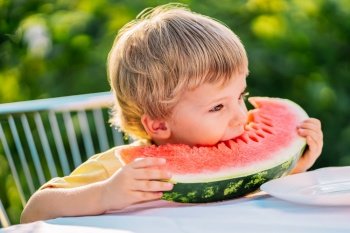 Handsome toddler boy eating fresh red watermelon. Yummy portrait of smiling child sits by the table in garden. Healthy lifestyle, melon vitamins, summer harvest. High quality photo. Handsome toddler boy eating fresh red watermelon.Yummy portrait of smiling child