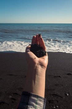 Close up woman holding pile of black sand on beach concept photo. First person view photography with sea waves on background. High quality picture for wallpaper, travel blog, magazine, article. Close up woman holding pile of black sand on beach concept photo