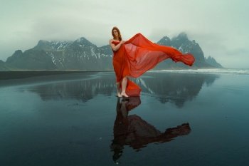 Beautiful woman in red dress on Reynisfjara beach scenic photography. Picture of person with hills on background. High quality wallpaper. Photo concept for ads, travel blog, magazine, article. Beautiful woman in red dress on Reynisfjara beach scenic photography