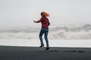 Funny tourist jumping on black Iceland beach scenic photography. Picture of person with stormy sea on background. High quality wallpaper. Photo concept for ads, travel blog, magazine, article. Funny tourist jumping on black Iceland beach scenic photography