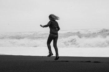 Joyful woman jumping on Reynisfjara beach monochrome scenic photography. Picture of person with stormy sea on background. High quality wallpaper. Photo concept for ads, travel blog, magazine, article. Joyful woman jumping on Reynisfjara beach monochrome scenic photography