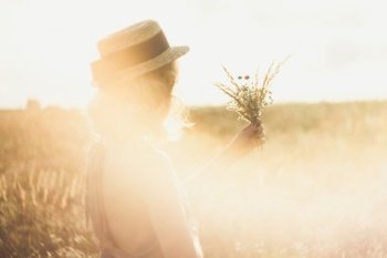 Close up girl in straw hat with bouquet surrounded by sunlight glare concept photo. Side view photography with meadow on background. High quality picture for wallpaper, travel blog, magazine, article. Close up girl in straw hat with bouquet surrounded by sunlight glare concept photo